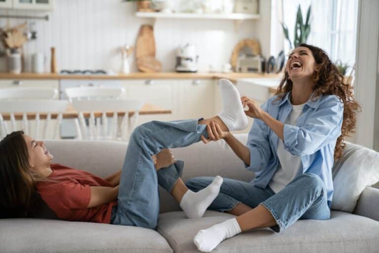 Overjoyed mother laughing out of loud while tickling feet of daughter, mom and teen girl having fun